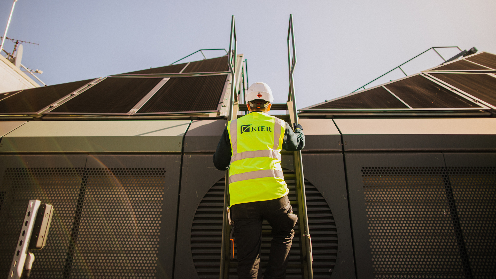 Kier worker climbing a ladder on a commercial building roof equipped with solar or thermal panels.